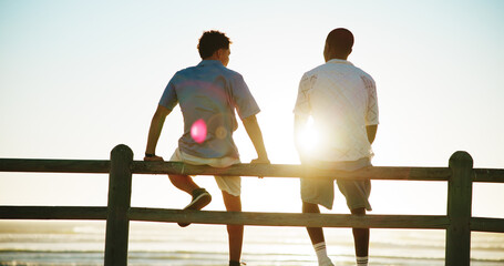 Men, sunset and back of friends at beach for break with bonding, thinking and support outdoor. Conversation, people and flare by wood fence, travel or vacation to relax with connection in nature