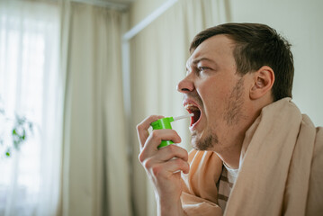 Fototapeta premium Sick man wrapped in a warm blanket, using throat spray to alleviate sore throat and flu symptoms while resting at home, seeking comfort and relief from illness