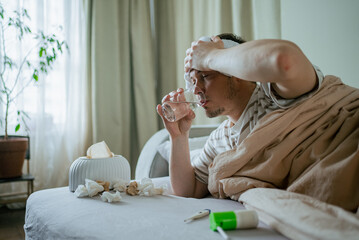 Sick man is lying on sofa under blanket, drinking water and holding wet towel on his forehead,...