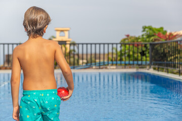 A boy in a swim trunks holding a red ball in a pool