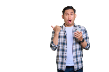 Shocked young Asian man in plaid shirt holding mobile phone and pointing thumbs aside at copy space isolated on transparent background
