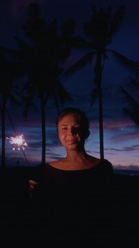 Woman holding sparkler. Christmas lights at the beach. Girl celebrate new year eve in tropical palm tree. Female and Bengal fire. Slow motion vertical video. Blue hour and coconut tree
