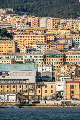 Obraz premium Genoa, Italy - September 22, 2024: Colorful hillside facades and layered historic buildings rise above the port waterfront with boats and greenery.