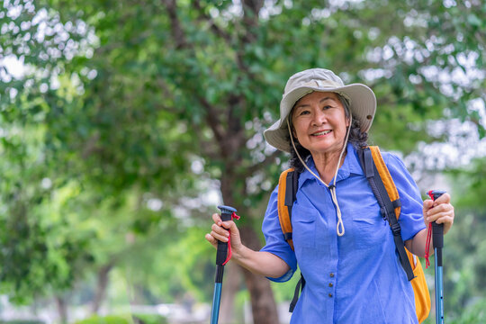 portrait cheerful senior woman wears hat,with backpack and trekking poles standing in nature,elderly asian female happy in hiking trip - Powered by Adobe