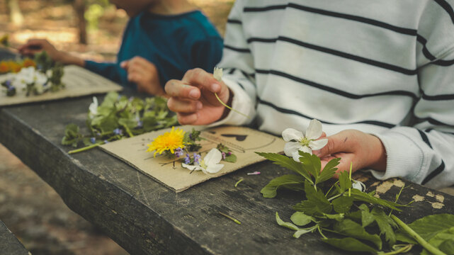 Children creating handmade flower crafts using cardboard, drawing vases and attaching real spring blossoms. Creative outdoor activity on a rustic wooden table in the forest