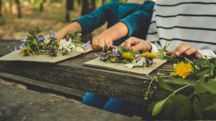 Children creating handmade flower crafts using cardboard, drawing vases and attaching real spring blossoms. Creative outdoor activity on a rustic wooden table in the forest