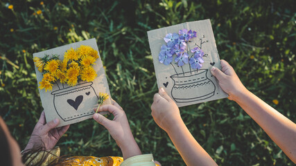 Two children creating flower crafts with fresh dandelions on cardboard, enjoying springtime nature and creative outdoor activity in a grassy field