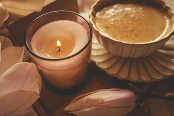 Lit candle in a glass jar with soft magnolia flowers, a gift box, and a coffee cup in warm pastel light on white lace fabric