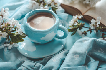 A cup of coffee with foam in a blue cup with white cloud design, surrounded by white spring blossoms, a soft blue fabric, and an open book, evoking a serene and cozy atmosphere