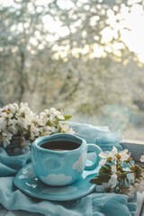 Blue cup with white clouds filled with tea or coffee, surrounded by cherry blossoms on a soft pastel fabric. Dreamy spring morning scene by the window