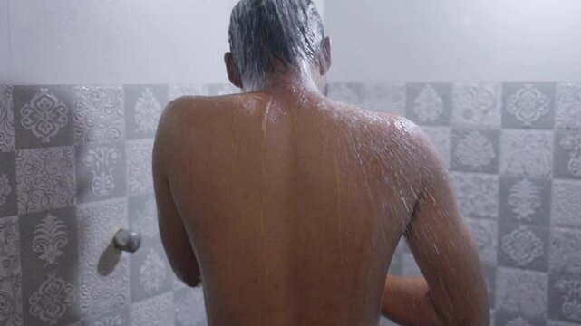 A backshot, slow-motion footage of a young man taking a hot shower in the bathroom during daytime. Healthcare and Hygiene concept