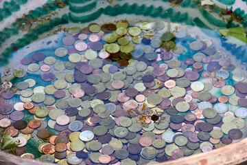 Coins scattered across a ceramic basin partially filled with water showing oxidized surfaces and varied patinas surrounded by faded green and white glaze detailing; aged coins;
