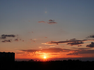 Amazing orange sunset over Kyiv city with buildings silhouettes in horizon. Urban cityscape of black buildings against the backdrop of glowing orb of the setting sun and red and lilac scenic clouds.