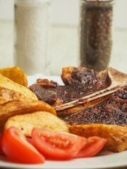 Cooked beef T bone steak meal. Premium high quality meat served on a plate with wedges and fresh tomato. Fine expensive well done meal.