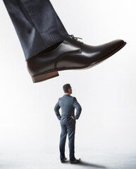 Tiny man office worker standing under pressure of the giant shoe of his big boss about to step on him. Workplace confrontation, leadership concept. Power dynamics or intimidation in business