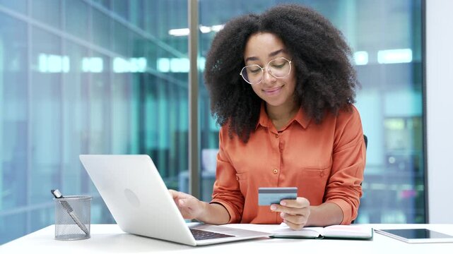 Happy african american businesswoman doing online shopping typing credit card data on laptop computer while sitting in business office. Smiling black woman makes purchase, enjoy e-commerce e-shopping