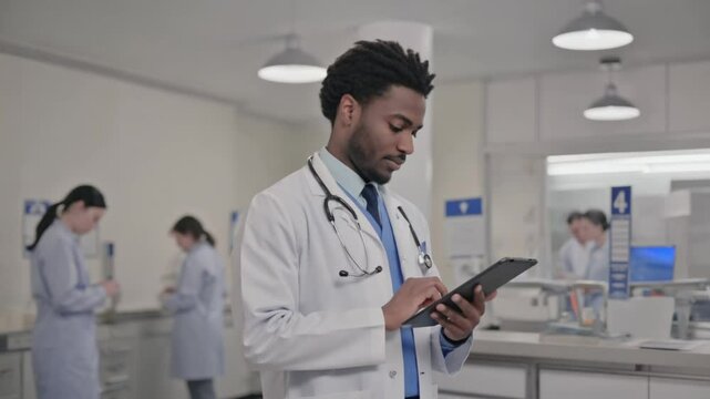An african american male physician taps notes on a tablet inside a busy clinical laboratory. Concept of innovative healthcare and real time data management.