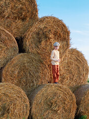 Portrait of child boy dressed in vintage style on background of round bales of straw, wheat harvest, yellow, orange, cap, copyspace, texture.
