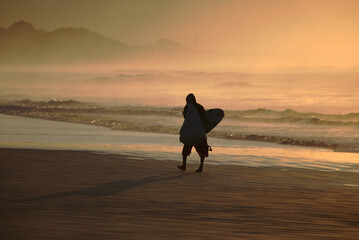 Surfer silhouette walking on the beach sand early morning before sunrise. Light reflections on sky and water. Itaguare beach, Bertioga, Brazil