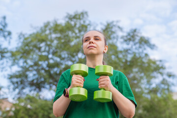 A woman in a green t-shirt performs a dumbbell shoulder press exercise during an outdoor workout...