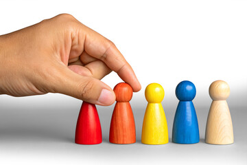 A close-up of two hands against a pastel green background. One hand is holding four colorful wooden human figurines (red, yellow, blue, and another natural wood tone), while the other hand is pointing