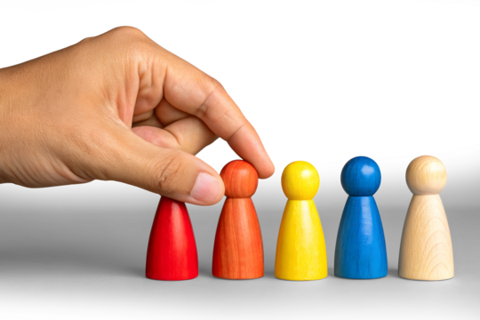 A close-up of two hands against a pastel green background. One hand is holding four colorful wooden human figurines (red, yellow, blue, and another natural wood tone), while the other hand is pointing