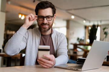 Businessman adjusting glasses while using smartphone and laptop in cafe