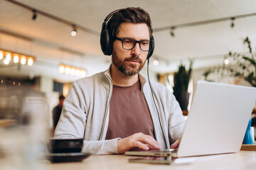 Fototapeta premium Focused businessman wearing headphones working on laptop in cafe