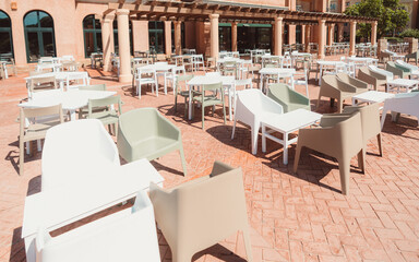 Outdoor terrace of a resort or restaurant with numerous plastic chairs and tables in soft neutral tones arranged on a patterned brick floor, in front of arched windows and columns, direct sunlight