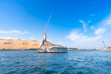 A felucca sailboat carrying tourists cruises along the Nile River near Aswan High Dam, Egypt, on a bright spring day with clear blue skies and light clouds.