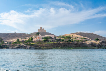 Aswan High Dam, Egypt: A scenic Nile River view in spring, featuring a historic monastery on a hill with greenery under a clear blue sky.
