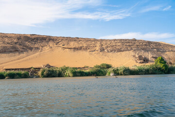 A scenic view of the Nile River flowing past sandy hills and vegetation near Aswan High Dam, Egypt, under a partly cloudy blue sky in springtime.