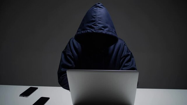 Anonymous hooded figure using laptop in dark setting, representing hacking and cybercrime

