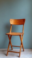 Wooden folding chair stands against a muted teal wall, resting on light flooring