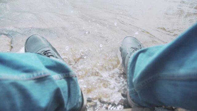 Close-up of the feet of an unemployed, sad man in boots on the river bank. Waves are hitting the boots.