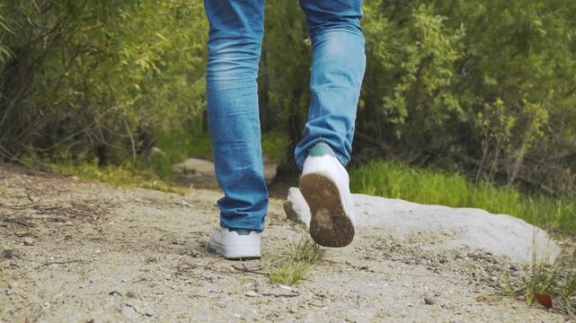 Head to toe close-up of a man in blue jeans and white sneakers walking from the beach into the woods.
