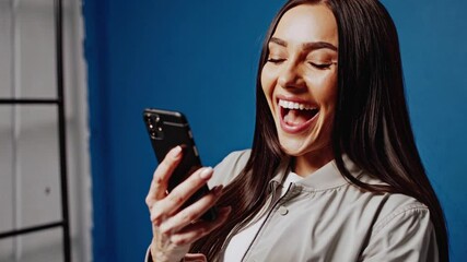 Smiling funny beautiful attractive young brunette woman 20s wearing basic jacket standing using mobile cell phone typing sms message isolated on bright blue colour wall background, studio portrait