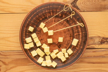 Fragrant hard cheese cut into cubes with bamboo skewers with a clay plate on a wooden table, macro, top view.