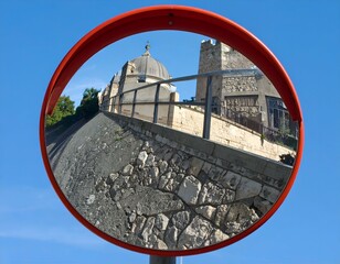 Convex mirror reflecting a historic building and stone wall under a clear blue sky.
