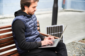 Man sits on wooden bench, working on laptop connected to solar panel. Urban setting and casual...