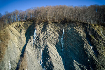 Rocky cliff with striking, layered striations, topped by dense forest of bare trees. Small frozen...