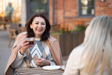 Middle aged woman sitting outdoor cafe, drinking coffee with friend and talking