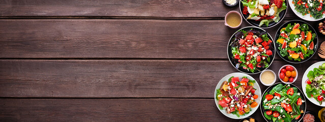 Assortment of delicious fresh salads. Top view side border on a dark wood banner background. Summer food concept.