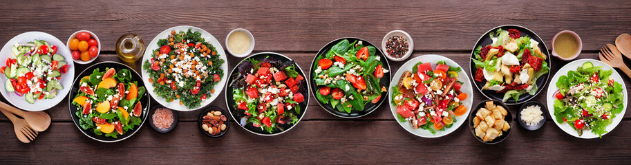 Variety of delicious fresh salads. Above view table scene on a dark wood banner background. Summer food concept.