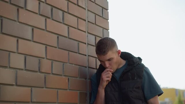 young male student wearing black vest and t-shirt rests against brick building wall inhaling vape device while coughing under daylight sky with faint background of city street and architecture