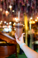 A hand holds a cold chocolate drink topped with syrup inside the Chocolate Museum café in Cartagena, Colombia, surrounded by elegant lighting and floral decor.