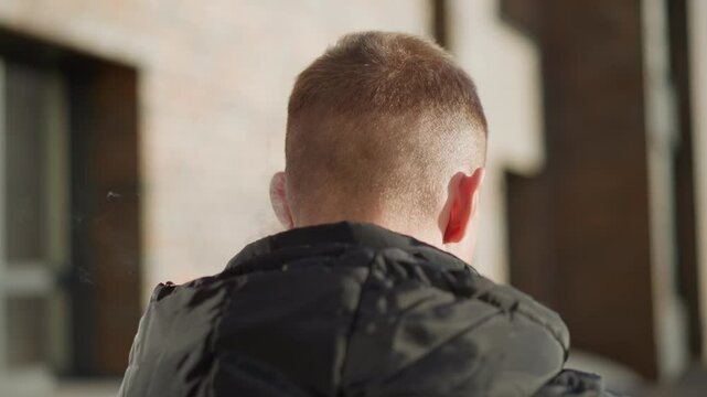 back view young man inhaling shisha outdoors on sunny day releasing thick smoke against blurred urban background with sunlight glow on jacket in vibrant warm late afternoon light soft haze