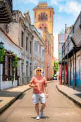 Young Latino man in coral shirt, white shorts, and straw hat confidently walks through a colonial...