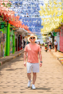 Young smiling Latino man walks along a decorated street in Getseman&iacute;, Cartagena, wearing coral shirt, white shorts, straw hat and sunglasses under vibrant papelillo banners.