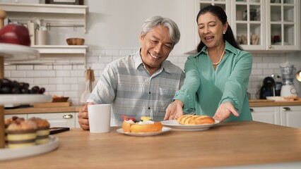 Smiling grandfather with casual cloth sitting at table while grandmother presenting breakfast. Senior couple talking and spending time or supporting together. Healthy relationship concept. Myrmidon.